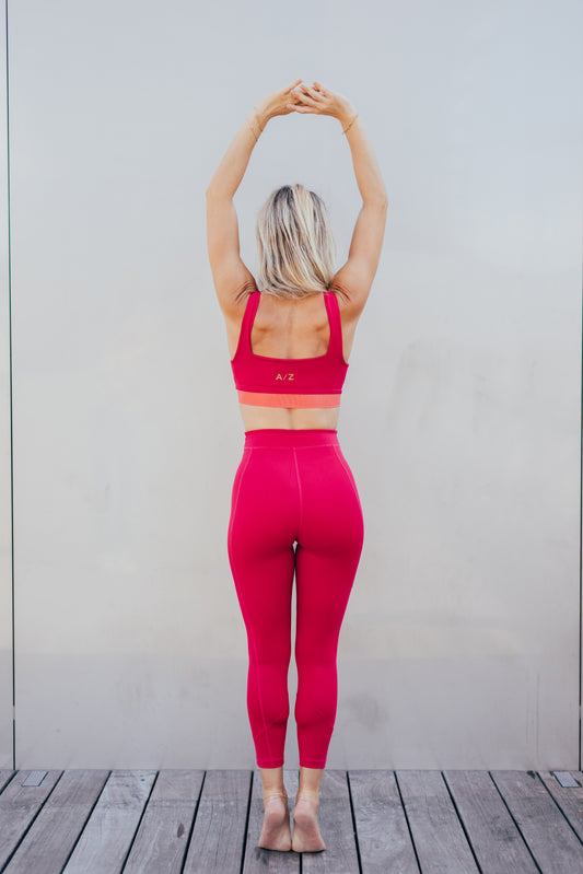 La coach sportive Julie Pujols posant debout avec les mains pointées en haut, de dos. Elle porte la tenue azar gang couleur framboise.