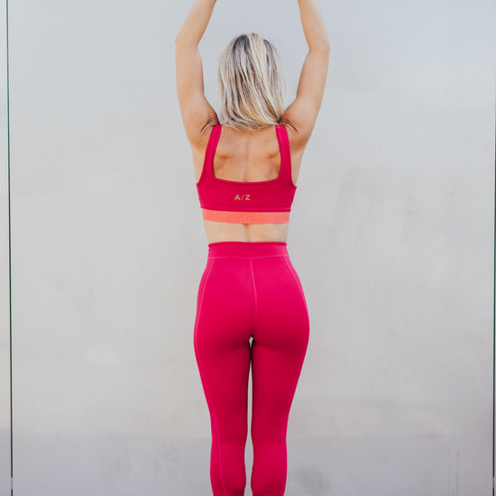La coach sportive Julie Pujols posant debout avec les mains pointées en haut, de dos. Elle porte la tenue azar gang couleur framboise.