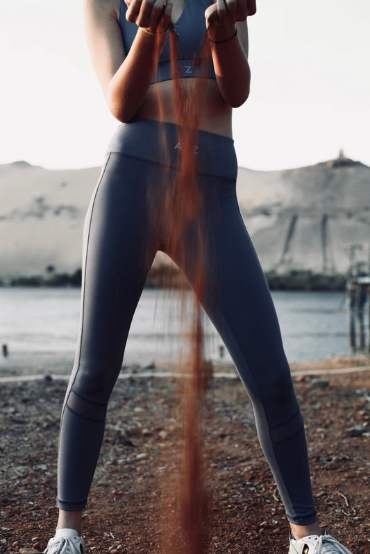 Femme debout faisant couler entre ses mains du sable. Elle porte une tenue de sports bleu/gris de la marque AZAR GANG